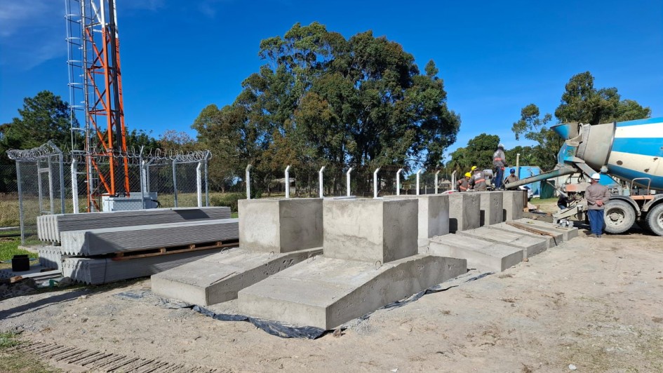 A foto mostra uma área de construção da obra da Penitenciária de Rio Grande. A fotografia mostra trabalhadores ao ar livre sob um céu azul claro. No centro, há várias fundações de concreto recém-construídas, algumas em forma de blocos retangulares e outras com uma base inclinada. À esquerda, pilhas de lajes de concreto pré-fabricadas estão em paletes de madeira, perto de uma cerca de arame e uma torre de telecomunicações alta, laranja e branca. No lado direito da imagem, um caminhão betoneira azul e branco está parado, e dois trabalhadores da construção civil com capacetes amarelos estão visíveis perto das fundações de concreto. O solo é de terra batida, com algumas trilhas de pneus. Árvores com folhagem verde densa formam o plano de fundo.