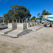 A foto mostra uma área de construção da obra da Penitenciária de Rio Grande. A fotografia mostra trabalhadores ao ar livre sob um céu azul claro. No centro, há várias fundações de concreto recém-construídas, algumas em forma de blocos retangulares e outras com uma base inclinada. À esquerda, pilhas de lajes de concreto pré-fabricadas estão em paletes de madeira, perto de uma cerca de arame e uma torre de telecomunicações alta, laranja e branca. No lado direito da imagem, um caminhão betoneira azul e branco está parado, e dois trabalhadores da construção civil com capacetes amarelos estão visíveis perto das fundações de concreto. O solo é de terra batida, com algumas trilhas de pneus. Árvores com folhagem verde densa formam o plano de fundo.