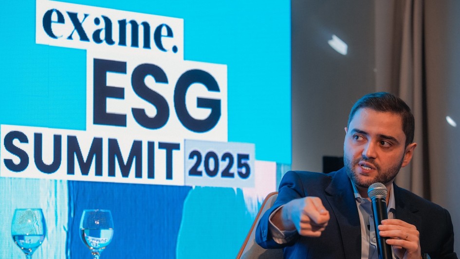 A imagem mostra o vice-governador do Estado, Gabriel Souza, em um painel de discussão. Ele é um homem branco de barba curta, vestindo um terno azul-escuro e camisa clara, sentado em uma poltrona cinza. Com uma mão, ele segura um microfone e com a outra, aponta para frente enquanto fala, com a boca ligeiramente aberta. Ao fundo, um telão azul exibe em branco o logotipo "exame." e as palavras "ESG SUMMIT 2025" em letras grandes. Dois copos de água vazios estão em uma mesa de vidro à frente do telão. Gabriel Souza foi um dos painelistas do evento ESG Summit 2025, que discute o futuro do clima e é promovido pela revista Exame, participando do painel "Eventos climáticos extremos e a necessidade de adaptação".