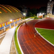 A foto noturna em alta definição mostra a pista de atletismo e o ginásio do Centro Estadual de Treinamento Esportivo (Cete) iluminados. A pista de corrida é vermelha escura, com algumas raias visíveis, e a área central é um campo de grama verde vibrante. Algumas pessoas, em tamanho pequeno devido à distância, estão correndo na pista. À esquerda, destaca-se a fachada de um grande ginásio com um telhado abobadado em tons de dourado e laranja devido à iluminação interna. As paredes do ginásio são azuis na parte inferior e brancas nos pilares. Postes de luz altos e brilhantes iluminam intensamente toda a área. Ao fundo, edifícios e árvores são visíveis sob o céu escuro.