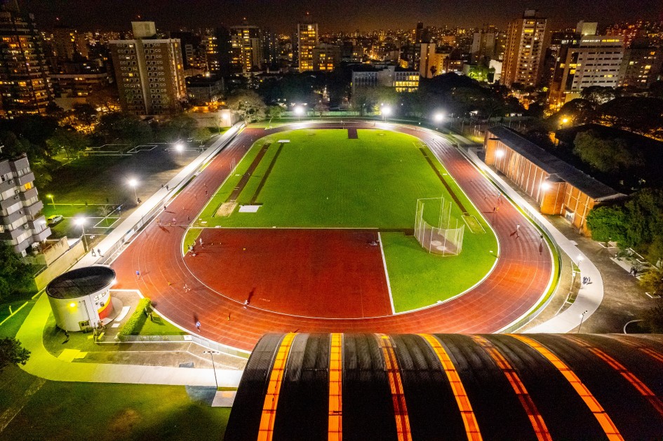 A foto noturna mostra uma vista aérea do Centro Estadual de Treinamento Esportivo (Cete). Uma pista de atletismo oval, com raias vermelhas e bem iluminada por postes de luz, domina o centro da imagem. Dentro da pista, há um campo de grama verde intenso. Várias pessoas pequenas podem ser vistas correndo na pista. No primeiro plano, à direita, aparece o telhado curvo de um ginásio, com luzes internas amareladas visíveis. Ao redor do complexo esportivo, à noite, destacam-se vários edifícios iluminados, sugerindo uma área urbana. A cidade de Porto Alegre aparece em segundo plano.
