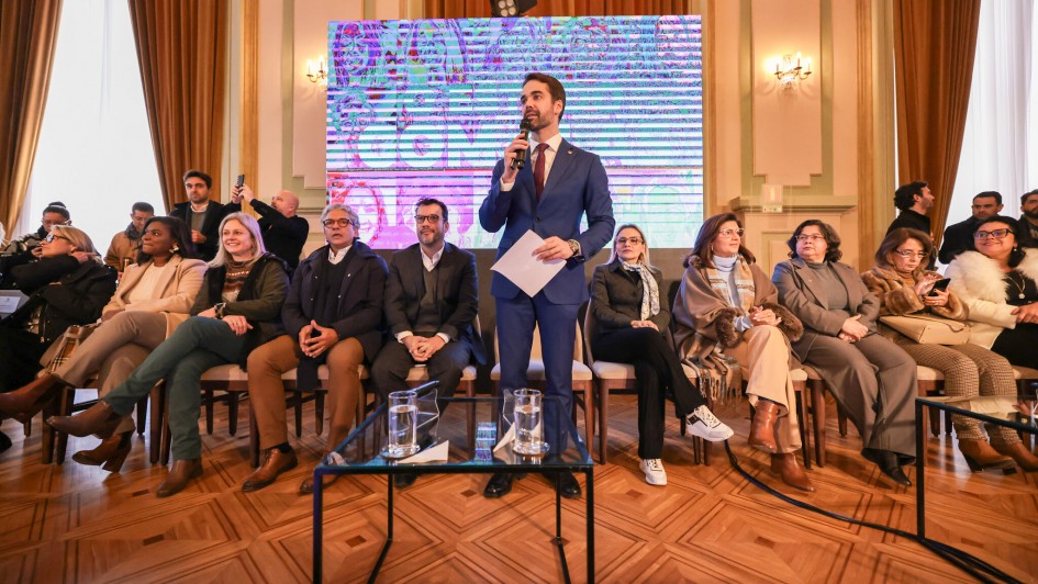 A imagem mostra o Governador Eduardo Leite, vestindo um terno azul escuro, camisa branca e gravata, discursando em um evento. Ele segura um microfone com a mão direita e papéis na esquerda. Atrás dele, há uma tela grande que exibe uma imagem distorcida ou abstrata em tons de roxo e azul. No palco, diversas pessoas estão sentadas em cadeiras de madeira com estofamento claro, observando o governador. Entre elas, há homens e mulheres de várias idades, alguns com óculos. A maioria das mulheres veste roupas de cores neutras ou escuras, como tons de marrom, cinza e bege. Um dos homens sentados na primeira fila está de terno escuro. O piso é de madeira, com um padrão de parquet. Há uma mesa de centro de vidro à frente do governador, com copos e uma jarra de água. O ambiente parece ser um salão, com cortinas claras e detalhes arquitetônicos clássicos ao fundo, indicando um local formal ou governamental.