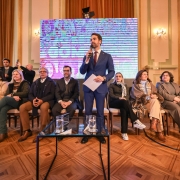 A imagem mostra o Governador Eduardo Leite, vestindo um terno azul escuro, camisa branca e gravata, discursando em um evento. Ele segura um microfone com a mão direita e papéis na esquerda. Atrás dele, há uma tela grande que exibe uma imagem distorcida ou abstrata em tons de roxo e azul. No palco, diversas pessoas estão sentadas em cadeiras de madeira com estofamento claro, observando o governador. Entre elas, há homens e mulheres de várias idades, alguns com óculos. A maioria das mulheres veste roupas de cores neutras ou escuras, como tons de marrom, cinza e bege. Um dos homens sentados na primeira fila está de terno escuro. O piso é de madeira, com um padrão de parquet. Há uma mesa de centro de vidro à frente do governador, com copos e uma jarra de água. O ambiente parece ser um salão, com cortinas claras e detalhes arquitetônicos clássicos ao fundo, indicando um local formal ou governamental.