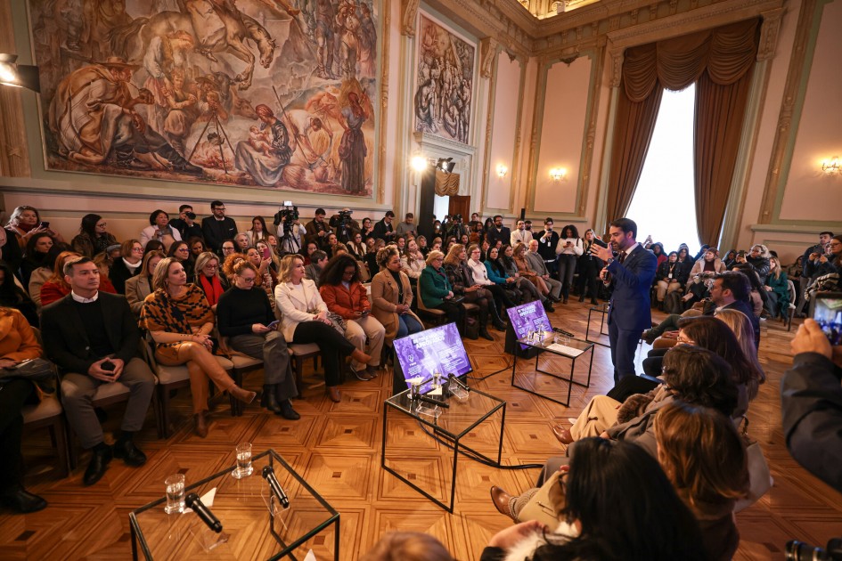 A imagem mostra o Governador Eduardo Leite, de terno escuro, discursando em um evento formal, segurando um microfone e olhando para a plateia. Ele está em pé, no lado direito da foto. O ambiente é um salão grande e ricamente decorado, com painéis de parede que apresentam murais artísticos detalhados, retratando cenas clássicas com cavalos e figuras humanas. Uma grande cortina pesada, de cor marrom ou bordô, adorna uma janela alta no lado direito da sala, com pregas elegantes. O teto é alto e ornamentado, e o chão possui um padrão de parquet. Uma vasta audiência, composta por homens e, em sua maioria, mulheres, está sentada em cadeiras dispostas em filas semicirculares. As pessoas na plateia estão atentas ao discurso do governador. Há mesas de centro de vidro com folhetos ou materiais promocionais, alguns com um design gráfico em tons de roxo, sugerindo o tema do evento. A iluminação é suave, complementando a atmosfera solene do local.