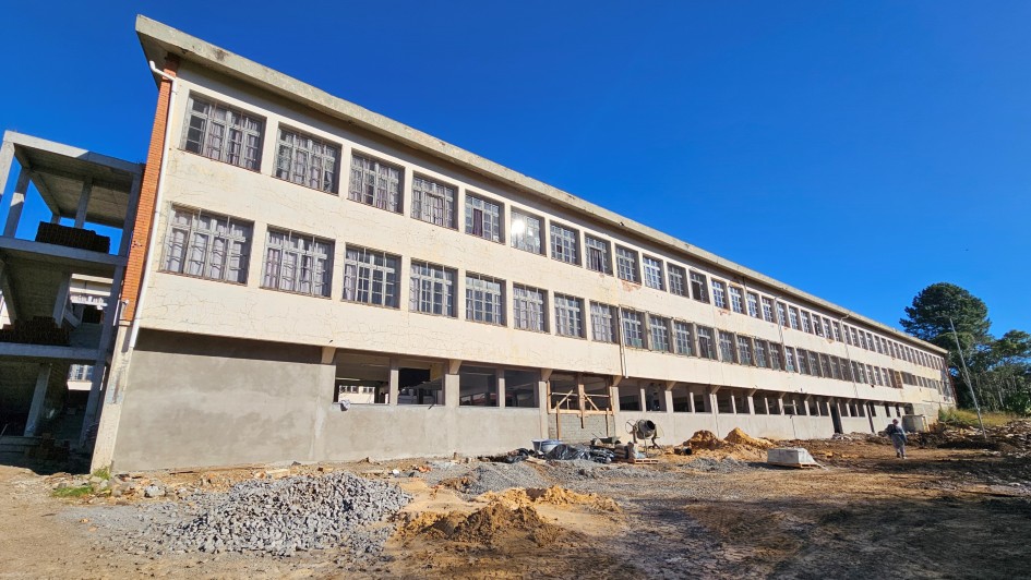 A imagem mostra um prédio de três andares em construção. A estrutura tem formato retangular e alongado, com várias janelas alinhadas horizontalmente em cada andar. O prédio está localizado em um terreno de terra batida, e ao redor dele há pilhas de materiais de construção espalhadas, indicando que a obra ainda está em andamento. O céu está claro e azul, com boa iluminação natural.