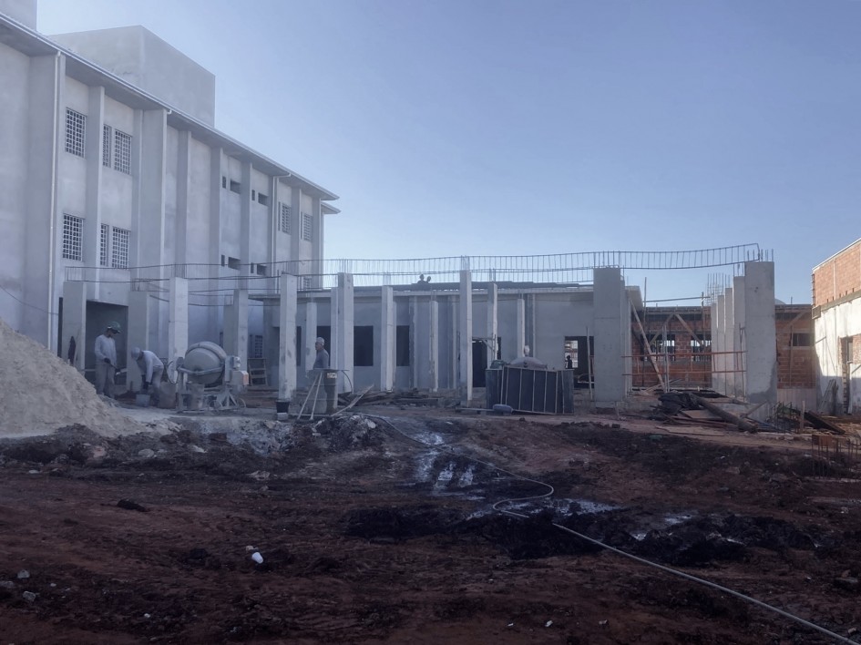 A imagem mostra um canteiro de obras ativo. No lado esquerdo, há um prédio de dois andares já construído, com janelas protegidas por grades. No centro e à direita da imagem, vê-se uma estrutura de concreto em construção, com colunas verticais e paredes parcialmente erguidas.
Há trabalhadores no local, envolvidos nas atividades da obra. Também estão visíveis equipamentos de construção, como uma betoneira (máquina usada para misturar concreto) e pilhas de materiais diversos. O chão está coberto por terra vermelha e há detritos da construção espalhados pelo terreno.