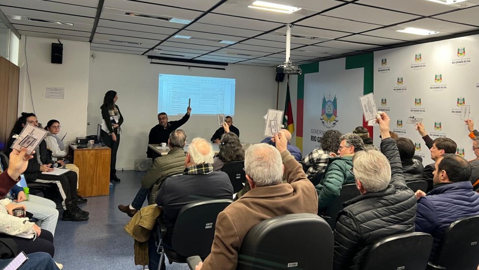 A imagem mostra uma sala com várias pessoas sentadas em cadeiras, todas voltadas para a parte da frente da sala. Na frente, há uma tela de projeção que exibe um documento ou planilha. Algumas pessoas na plateia estão levantando papéis, embora o conteúdo exato não seja visível. Na frente da sala, há três pessoas: uma está em pé e as outras duas estão sentadas atrás de uma mesa. Ao fundo, na parede, há um logotipo do "Governo do Rio Grande do Sul".