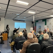A imagem mostra uma sala com várias pessoas sentadas em cadeiras, todas voltadas para a parte da frente da sala. Na frente, há uma tela de projeção que exibe um documento ou planilha. Algumas pessoas na plateia estão levantando papéis, embora o conteúdo exato não seja visível. Na frente da sala, há três pessoas: uma está em pé e as outras duas estão sentadas atrás de uma mesa. Ao fundo, na parede, há um logotipo do "Governo do Rio Grande do Sul".