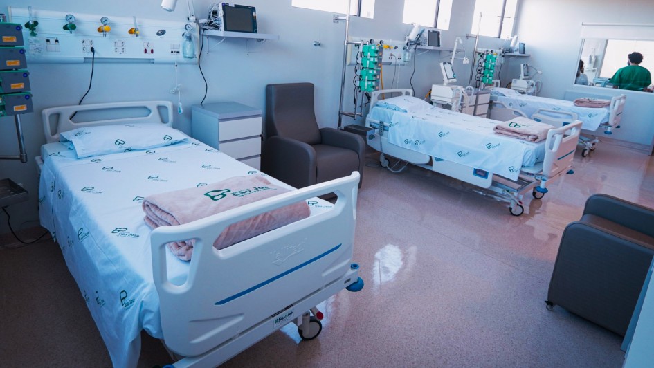 A imagem mostra um quarto de hospital com três leitos equipados. Cada leito possui: uma cama hospitalar ajustável, coberta com lençóis brancos que exibem o logotipo do hospital; equipamentos médicos ao lado, como monitores e bombas de infusão, e cadeiras reclináveis próximas às camas, provavelmente destinadas a acompanhantes ou visitantes. Ao fundo, há janelas que permitem a entrada de luz natural, iluminando o ambiente. No canto direito da imagem, aparece uma pessoa vestida com roupas verdes, típicas de profissionais da saúde.
