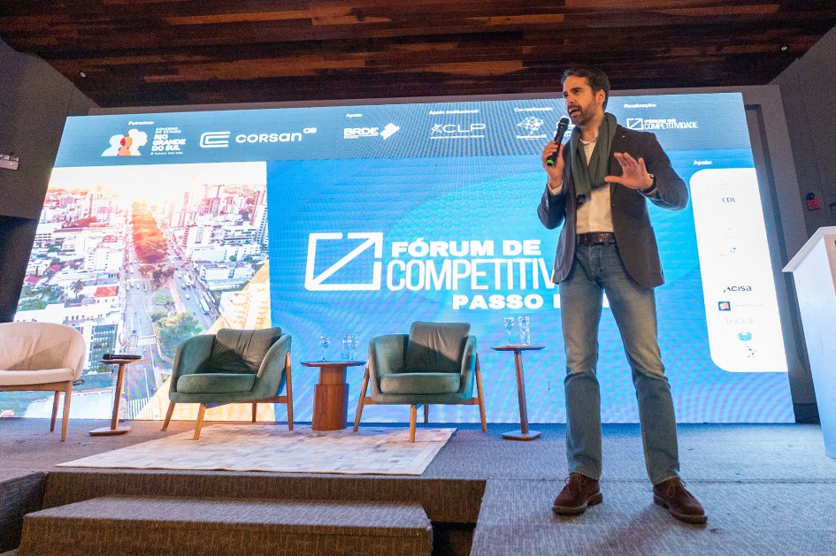 Na imagem, o governador Eduardo Leite aparece em pé no palco durante o Fórum de Competitividade, realizado em Passo Fundo. Ele segura um microfone e está em meio a um discurso ou apresentação, com expressão séria e postura confiante. Ao fundo, um telão exibe o nome do evento e os logotipos de apoiadores como Corsan, BRDE e CLP. O ambiente é formal e voltado ao debate sobre desenvolvimento e infraestrutura, reforçando o papel do governador como protagonista nas estratégias para impulsionar o norte do Rio Grande do Sul.