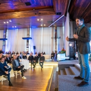 Na imagem, o governador Eduardo Leite aparece discursando em um evento realizado em um salão amplo e bem iluminado, com piso e teto de madeira. Ele está de pé, segurando um microfone, e se dirige a uma plateia atenta, sentada em mesas redondas. O ambiente é elegante, com iluminação azul e janelas altas com cortinas brancas. Ao fundo, há um telão exibindo logotipos, incluindo o da Corsan, sinalizando a formalidade e importância institucional do encontro.