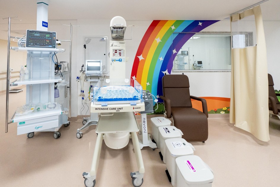 A imagem mostra um quarto hospitalar em uma unidade de terapia intensiva. No centro da imagem, há uma incubadora com a inscrição “INTENSIVE CARE UNIT” (Unidade de Terapia Intensiva), equipada com diversos aparelhos médicos conectados. À esquerda, há um equipamento médico alto, com monitores e prateleiras contendo suprimentos hospitalares. À direita, vê-se uma poltrona reclinável marrom. Ao fundo, a parede é decorada com um mural colorido de arco-íris e estrelas, trazendo um toque lúdico ao ambiente clínico.