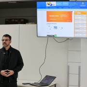 A imagem mostra uma pessoa em pé ao lado de uma mesa, sobre a qual há um laptop aberto. Acima da pessoa, há uma tela grande exibindo um site do programa Nota Fiscal Gaúcha. À direita da imagem, há um banner vertical da Aapecan (Associação de Apoio a Pessoas com Câncer). O banner contém o lema "ATENDER • ORIENTAR • INTEGRAR" e um emblema com mãos dadas dentro de um círculo, simbolizando apoio e solidariedade.