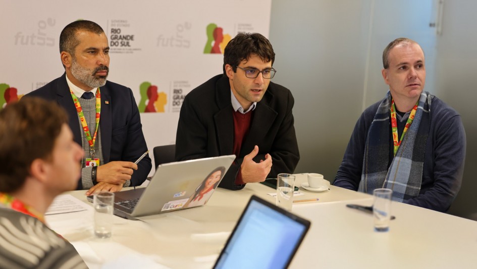 A foto mostra três homens adultos sentados ao redor de uma mesa de reunião branca, com laptops, copos de água e xícaras de café. O homem do centro, de óculos e casaco escuro sobre um suéter vermelho, gesticula enquanto fala, sendo o foco principal da imagem. À sua esquerda, um homem com barba grisalha, terno e suéter, ouve atentamente. À direita, outro homem, usando um cachecol xadrez azul, também está atento. Um quarto homem, de costas para a câmera, é parcialmente visível no canto inferior esquerdo. Ao fundo, um banner branco com logotipos do Governo do Estado do Rio Grande do Sul em vermelho e verde, e a palavra "futsal" repetida em tons de cinza claro. A cena sugere uma discussão ou apresentação profissional.