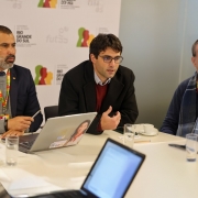 A foto mostra três homens adultos sentados ao redor de uma mesa de reunião branca, com laptops, copos de água e xícaras de café. O homem do centro, de óculos e casaco escuro sobre um suéter vermelho, gesticula enquanto fala, sendo o foco principal da imagem. À sua esquerda, um homem com barba grisalha, terno e suéter, ouve atentamente. À direita, outro homem, usando um cachecol xadrez azul, também está atento. Um quarto homem, de costas para a câmera, é parcialmente visível no canto inferior esquerdo. Ao fundo, um banner branco com logotipos do Governo do Estado do Rio Grande do Sul em vermelho e verde, e a palavra "futsal" repetida em tons de cinza claro. A cena sugere uma discussão ou apresentação profissional.