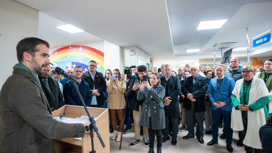 A imagem mostra um grupo de pessoas reunidas em um hospital. Em destaque, o governador Eduardo Leite está falando em um púlpito com microfones, dirigindo-se ao público presente. Ao fundo, há um mural colorido com um arco-íris pintado na parede, o que adiciona um toque acolhedor ao ambiente hospitalar.