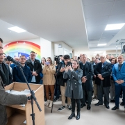 A imagem mostra um grupo de pessoas reunidas em um hospital. Em destaque, o governador Eduardo Leite está falando em um púlpito com microfones, dirigindo-se ao público presente. Ao fundo, há um mural colorido com um arco-íris pintado na parede, o que adiciona um toque acolhedor ao ambiente hospitalar.