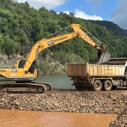 A imagem mostra uma escavadeira Hyundai 260LC-9 carregando pedras em um caminhão basculante, em uma área próxima a um rio. O solo é composto por pedras e cascalho, e ao fundo há vegetação densa. A cena representa uma etapa de desassoreamento ou intervenção de infraestrutura para controle de enchentes ou recuperação ambiental.