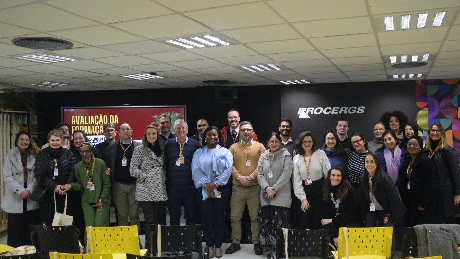 A foto mostra um grande grupo de pessoas, aproximadamente 30, de diversas etnias e idades, em pé e posando para uma foto em um ambiente interno. A maioria está sorrindo e olhando para a câmera. Ao fundo, uma parede escura com o logotipo "PROCERGS" em branco e designs coloridos no lado direito. À esquerda, parte de um banner vermelho com os dizeres "AVALIAÇÃO DA FORMAÇÃO" é visível. O teto possui diversas luminárias retangulares e circulares. No primeiro plano, há fileiras de cadeiras amarelas vazias. As pessoas estão vestindo uma variedade de roupas casuais e sociais, e algumas usam crachás no pescoço.