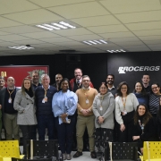 A foto mostra um grande grupo de pessoas, aproximadamente 30, de diversas etnias e idades, em pé e posando para uma foto em um ambiente interno. A maioria está sorrindo e olhando para a câmera. Ao fundo, uma parede escura com o logotipo "PROCERGS" em branco e designs coloridos no lado direito. À esquerda, parte de um banner vermelho com os dizeres "AVALIAÇÃO DA FORMAÇÃO" é visível. O teto possui diversas luminárias retangulares e circulares. No primeiro plano, há fileiras de cadeiras amarelas vazias. As pessoas estão vestindo uma variedade de roupas casuais e sociais, e algumas usam crachás no pescoço.
