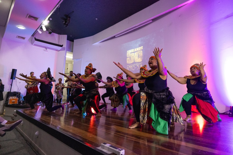 A foto captura uma performance de dança afro-brasileira do grupo Odomodê em um palco de madeira, iluminado por luzes roxas e rosadas. Cerca de nove dançarinos, a maioria mulheres e alguns homens, estão em diferentes posições. Muitos estão com os braços estendidos e palmas das mãos abertas para cima, com as pernas dobradas, em uma postura que evoca força e conexão. Eles vestem figurinos coloridos, com detalhes que incluem tecidos estampados, contas e cocares estilizados. Ao fundo, um telão branco exibe o texto "50 ANOS", parcialmente visível, e "AFRO-SUL". A cena é dinâmica e cheia de movimento.