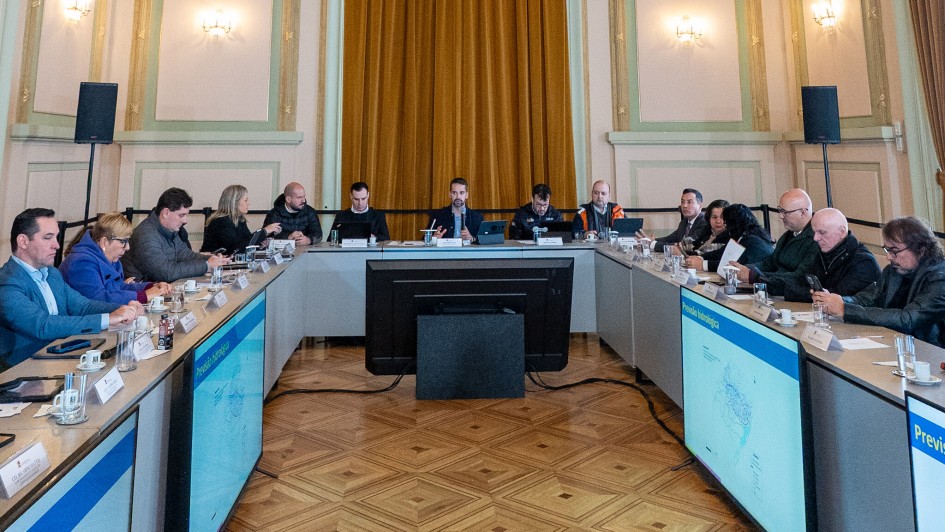 A imagem mostra uma sala de conferências com várias pessoas sentadas ao redor de uma mesa em formato de U, com o governador ao centro. Em frente a cada participante, há plaquinhas com nomes, microfones e documentos, sugerindo uma reunião formal ou institucional. Em ambos os lados da mesa, há duas telas grandes exibindo informações meteorológicas.