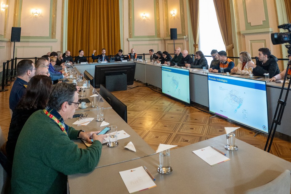 A imagem mostra uma sala de conferências com várias pessoas sentadas ao redor de uma grande mesa em formato de U. Sobre a mesa, há copos de água, papéis e microfones. Na sala, destacam-se dois grandes monitores que exibem mapas e textos com o título “Previsão Hidrológica”.