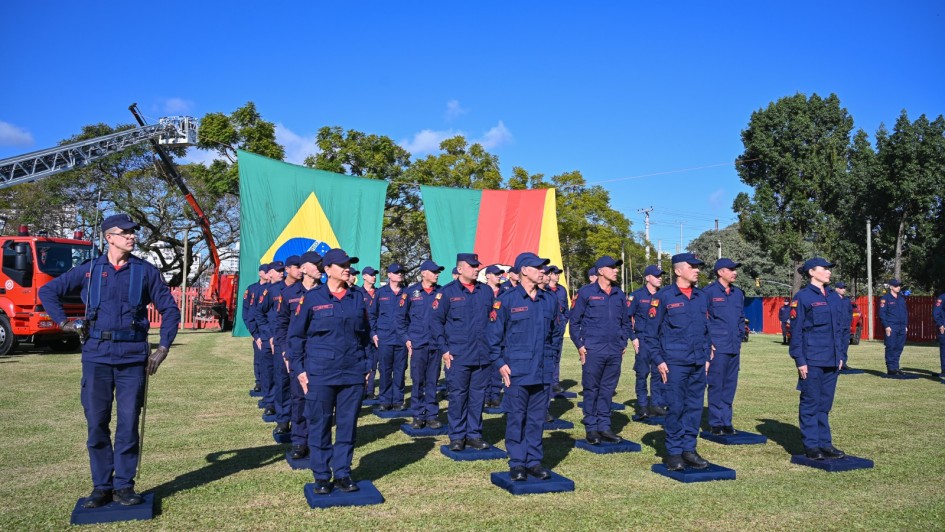 A imagem retrata uma cerimônia de formatura do Corpo de Bombeiros Militar do Rio Grande do Sul. Bombeiros uniformizados estão perfilados em um gramado, com bandeiras do Brasil e do RS ao fundo, além de um caminhão e equipamentos de resgate visíveis. O cenário é ao ar livre, com céu limpo e árvores ao redor, transmitindo um clima solene e de celebração da conquista profissional.