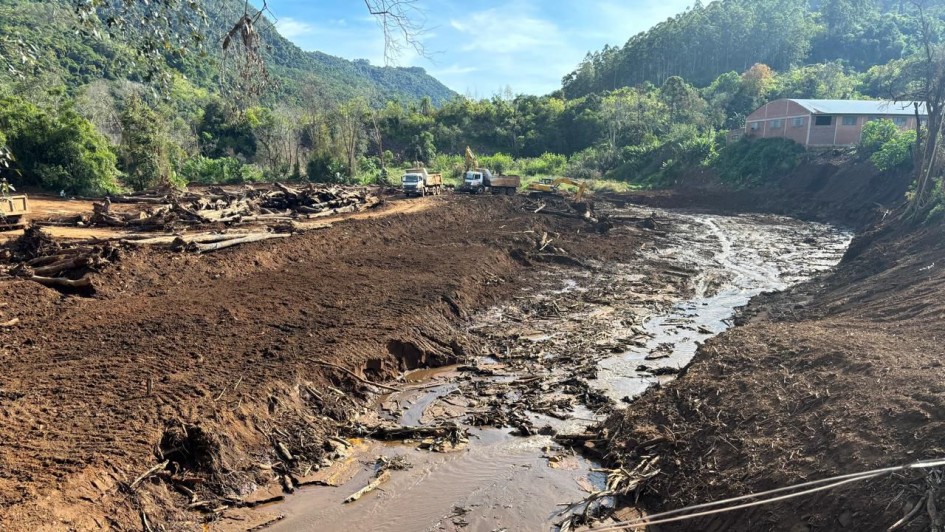 A imagem mostra uma área em Muçum que passou por grande destruição, provavelmente causada por enchente ou deslizamento. O solo está coberto por lama e entulhos, com troncos de árvores espalhados. Equipamentos pesados, como escavadeiras e caminhões, trabalham na limpeza do local, evidenciando um esforço de reconstrução em meio a um cenário de desastre natural. Ao fundo, há vegetação e edificações que ajudam a contextualizar a área atingida.