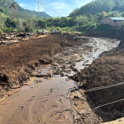 A imagem mostra uma área em Muçum que passou por grande destruição, provavelmente causada por enchente ou deslizamento. O solo está coberto por lama e entulhos, com troncos de árvores espalhados. Equipamentos pesados, como escavadeiras e caminhões, trabalham na limpeza do local, evidenciando um esforço de reconstrução em meio a um cenário de desastre natural. Ao fundo, há vegetação e edificações que ajudam a contextualizar a área atingida.