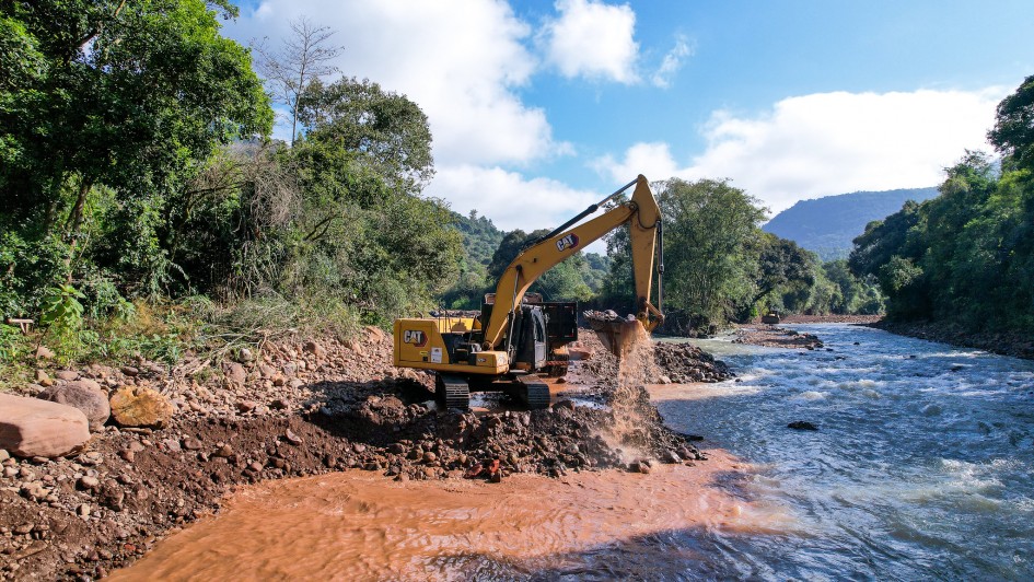 A imagem mostra uma escavadeira operando na margem de um rio. A máquina está removendo sedimentos da beira do rio, o que faz com que a água próxima fique turbulenta e barrenta. Ao redor, a paisagem é rica em vegetação verde, com árvores densas e mato cobrindo o terreno. 