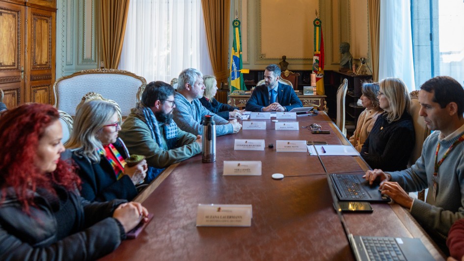 Imagem mostra mesa com 9 pessoas sentadas em cadeiras ao redor, cinco de um lado e três de outro. Na ponta, no centro da imagem, uma pessoa parece ouvir as outras. Sobre a mesa aparecem placas com nomes escritos. Uma das pessoas digita em um computador. Ao fundo, as bandeiras do Brasil e do Rio Grande do Sul