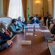 Imagem mostra mesa com 9 pessoas sentadas em cadeiras ao redor, cinco de um lado e três de outro. Na ponta, no centro da imagem, uma pessoa parece ouvir as outras. Sobre a mesa aparecem placas com nomes escritos. Uma das pessoas digita em um computador. Ao fundo, as bandeiras do Brasil e do Rio Grande do Sul
