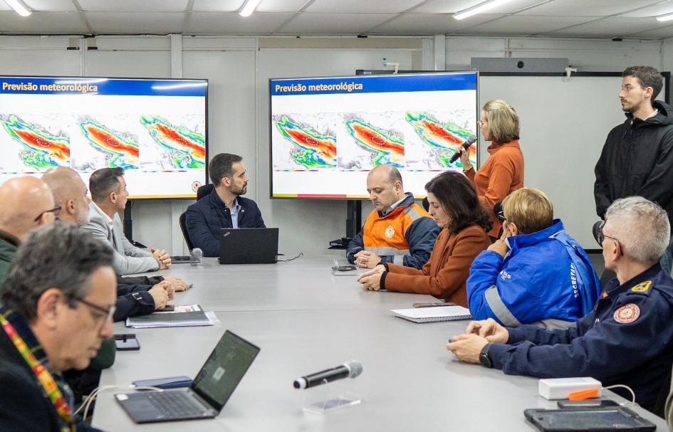 A imagem mostra o governador Eduardo Leite reunido com prefeitos e autoridades estaduais em uma sala de reuniões equipada com telas exibindo mapas meteorológicos. O encontro, realizado em 27 de junho de 2025, teve como foco o alinhamento de estratégias para enfrentar um novo evento climático extremo previsto para o Rio Grande do Sul. A cena transmite um momento de coordenação técnica e preventiva diante da ameaça de chuvas intensas.