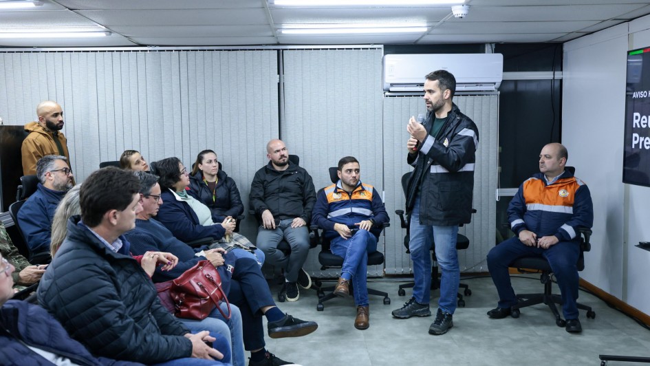 A foto mostra o governador Eduardo Leite (ao centro, em pé, de casaco escuro) conversando com um grupo de pessoas sentadas em uma sala. Ele segura um objeto pequeno em uma das mãos enquanto gesticula com a outra, parecendo explicar algo. As pessoas sentadas, algumas vestindo coletes da Defesa Civil, o observam atentamente. A sala parece ser um ambiente de escritório ou reunião, com persianas e ar condicionado. No canto superior direito, há um monitor exibindo parte de um texto.