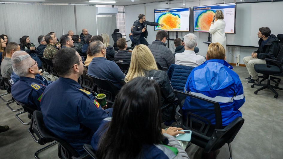 A foto mostra uma reunião em um auditório com teto de luminárias fluorescentes. Ao centro, de costas para a câmera, uma plateia diversa senta em cadeiras pretas. À frente, em um palco baixo, o governador Eduardo Leite, vestindo um casaco preto, aponta para um dos dois telões brancos. Os telões exibem mapas de calor com previsões meteorológicas em tons de laranja, azul e amarelo. Ao lado dele, à direita, uma mulher de casaco branco e um homem de moletom escuro também observam as telas. Um projetor está visível no teto. A cena retrata uma reunião de crise para avaliar o rescaldo de um temporal, com foco em dados meteorológicos.