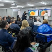 A foto mostra uma reunião em um auditório com teto de luminárias fluorescentes. Ao centro, de costas para a câmera, uma plateia diversa senta em cadeiras pretas. À frente, em um palco baixo, o governador Eduardo Leite, vestindo um casaco preto, aponta para um dos dois telões brancos. Os telões exibem mapas de calor com previsões meteorológicas em tons de laranja, azul e amarelo. Ao lado dele, à direita, uma mulher de casaco branco e um homem de moletom escuro também observam as telas. Um projetor está visível no teto. A cena retrata uma reunião de crise para avaliar o rescaldo de um temporal, com foco em dados meteorológicos.
