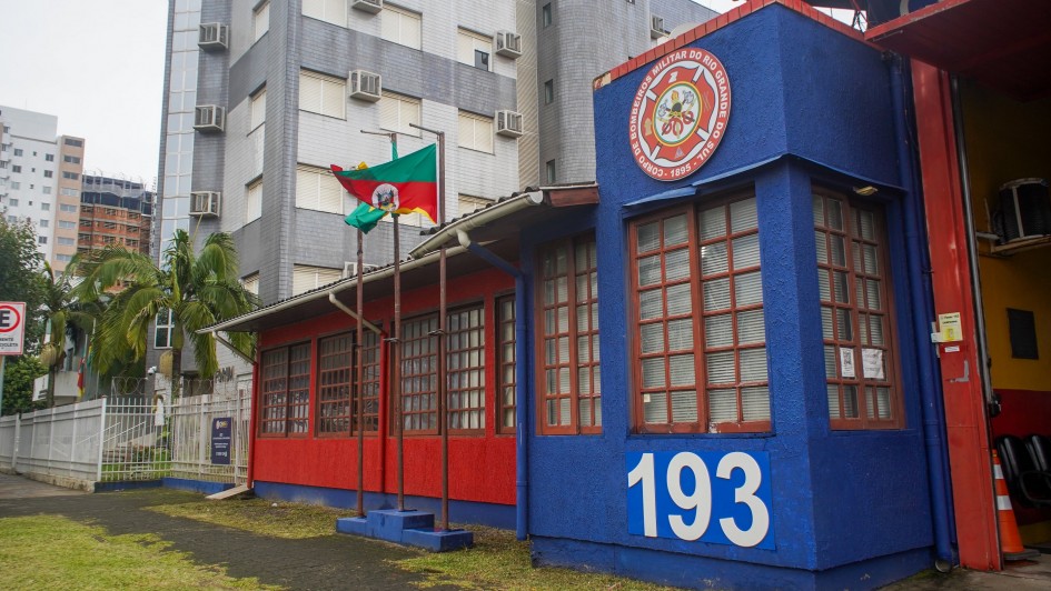 A foto mostra o prédio do Corpo de Bombeiros de Torres, com a frente pintada em azul vibrante e vermelho. A parte azul, à direita, exibe o número "193" em grandes algarismos brancos, e acima, um emblema circular detalhado com o nome "CORPO DE BOMBEIROS MILITAR DO RIO GRANDE DO SUL". A parte vermelha, à esquerda, tem janelas retangulares de esquadrias marrons. Uma bandeira do Rio Grande do Sul, com suas cores verde, vermelho e amarelo, tremula em um mastro no centro da imagem. Palmeiras e outras árvores verdes são visíveis. A estrutura foi instalada pelo governo do Estado e coordenada pela Defesa Civil estadual.