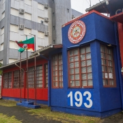 A foto mostra o prédio do Corpo de Bombeiros de Torres, com a frente pintada em azul vibrante e vermelho. A parte azul, à direita, exibe o número "193" em grandes algarismos brancos, e acima, um emblema circular detalhado com o nome "CORPO DE BOMBEIROS MILITAR DO RIO GRANDE DO SUL". A parte vermelha, à esquerda, tem janelas retangulares de esquadrias marrons. Uma bandeira do Rio Grande do Sul, com suas cores verde, vermelho e amarelo, tremula em um mastro no centro da imagem. Palmeiras e outras árvores verdes são visíveis. A estrutura foi instalada pelo governo do Estado e coordenada pela Defesa Civil estadual.