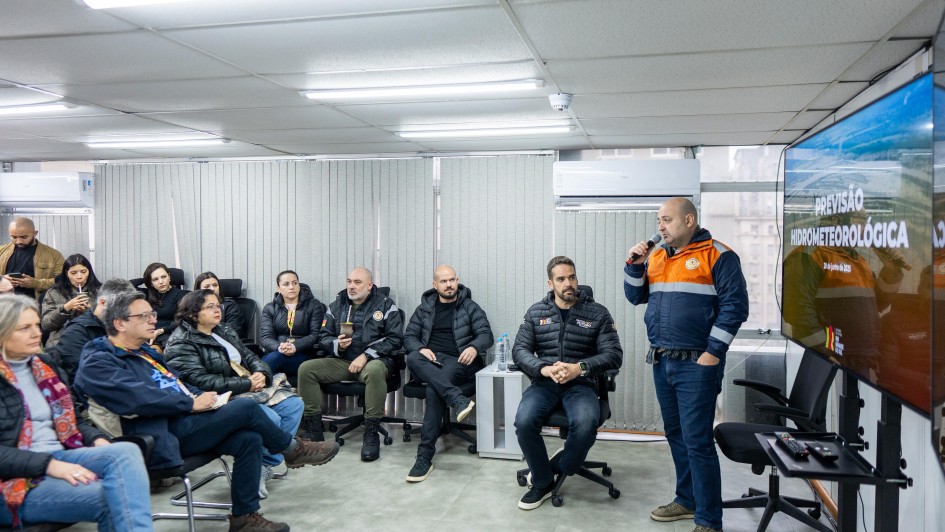 A imagem mostra um grupo de pessoas sentadas em uma sala, participando de uma apresentação. Uma pessoa está em pé, falando ao microfone, enquanto as demais estão atentas, ouvindo. O governador está sentado ao centro. Ao fundo, há uma tela grande de projeção exibindo o título "Apresentação hidrometeorológica",