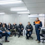 A imagem mostra um grupo de pessoas sentadas em uma sala, participando de uma apresentação. Uma pessoa está em pé, falando ao microfone, enquanto as demais estão atentas, ouvindo. O governador está sentado ao centro. Ao fundo, há uma tela grande de projeção exibindo o título "Apresentação hidrometeorológica",