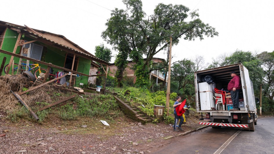 A imagem mostra uma cena à beira de uma estrada, onde duas pessoas estão carregando itens na parte traseira de um caminhão. O caminhão está estacionado à direita da imagem. Uma pessoa está dentro do caminhão, enquanto a outra está do lado de fora, alcançando os itens que estão sendo carregados.