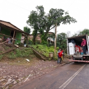 A imagem mostra uma cena à beira de uma estrada, onde duas pessoas estão carregando itens na parte traseira de um caminhão. O caminhão está estacionado à direita da imagem. Uma pessoa está dentro do caminhão, enquanto a outra está do lado de fora, alcançando os itens que estão sendo carregados.