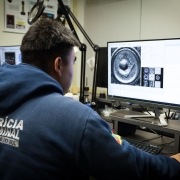 
A foto mostra um perito criminal, visto de costas, sentado em uma mesa de trabalho e focado em um monitor de computador. Ele veste um moletom azul escuro com os dizeres "PERÍCIA CRIMINAL RIO GRANDE DO SUL" em letras brancas na parte de trás, e o brasão do estado na manga direita. Na tela principal, uma imagem detalhada de um projétil ou cápsula de munição é ampliada, juntamente com outras imagens menores em miniatura. À esquerda do perito, há outro monitor e, ao fundo, um laboratório com equipamentos e uma prateleira com itens. A cena indica um trabalho de análise forense.