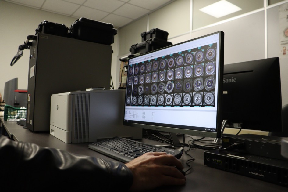 A foto, tirada de perto, mostra a mão direita de um perito criminal operando um mouse de computador. Ele usa uma jaqueta de couro preta e está sentado em frente a dois monitores. O monitor maior, à direita, exibe múltiplas imagens em miniatura de projéteis ou cápsulas de munição, organizadas em uma grade, com uma imagem ampliada no canto superior esquerdo. No lado esquerdo da mesa, há uma impressora e outros equipamentos de laboratório. O ambiente é um laboratório, com uma parede clara e prateleiras ao fundo. A cena destaca a análise detalhada de evidências balísticas.