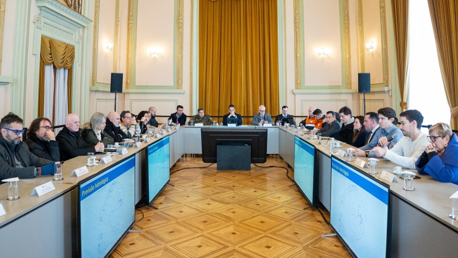 A imagem mostra uma reunião ocorrendo em uma sala elegante. Os participantes estão sentados ao redor de uma mesa em formato de U, com plaquinhas de identificação e microfones à frente de cada um. Ao centro da imagem, está o governador Eduardo Leite. 