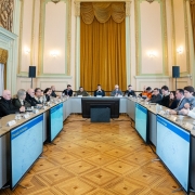 A imagem mostra uma reunião ocorrendo em uma sala elegante. Os participantes estão sentados ao redor de uma mesa em formato de U, com plaquinhas de identificação e microfones à frente de cada um. Ao centro da imagem, está o governador Eduardo Leite. 