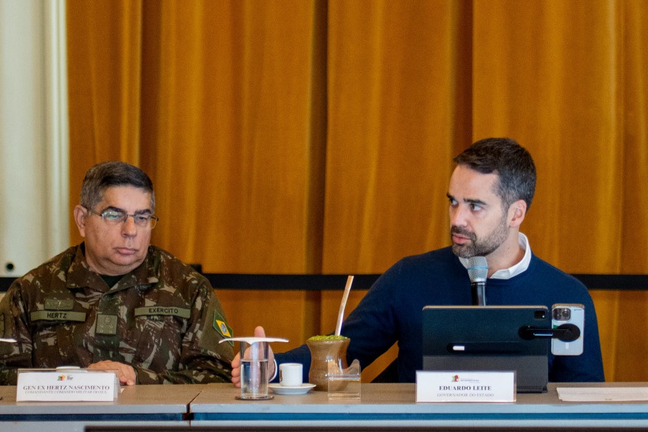 A imagem mostra duas pessoas sentadas à mesa, ambas com microfones à frente, participando de uma reunião. À direita, está o governador Eduardo Leite. À esquerda imagem, está o comandante Militar do Sul, general Hertz.