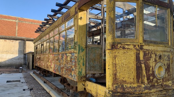 A imagem mostra um bonde antigo de cor amarela, posicionado sobre trilhos. O veículo está em estado de deterioração, com tinta descascada e ferrugem visível em várias partes da estrutura. Apesar disso, o formato original do bonde ainda é reconhecível, e a maioria das janelas permanece intacta.
