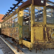 A imagem mostra um bonde antigo de cor amarela, posicionado sobre trilhos. O veículo está em estado de deterioração, com tinta descascada e ferrugem visível em várias partes da estrutura. Apesar disso, o formato original do bonde ainda é reconhecível, e a maioria das janelas permanece intacta.